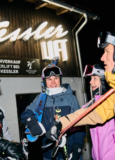 Vier Skifahrer in Winterkleidung mit Helmen und Skiern, fröhlich im Après-Ski bei Nacht im Kleinwalsertal, im Hintergrund leuchtet ein Neon-Schild. | © Kleinwalsertal Tourismus eGen • Fotograf: Oliver Farys
