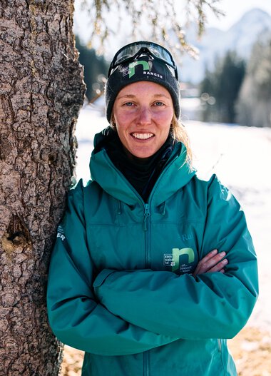 Porträt einer Frau im Winter, lachend mit verschränkten Armen, neben einem Baum, im Hintergrund Schnee und Berge. | © Kleinwalsertal Tourismus eGen • Fotograf: Oliver Farys