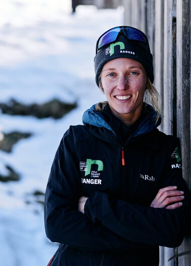 Frau in Winterkleidung mit Sonnenbrille und Mütze steht im Schnee neben einer Holzlounge. | © Kleinwalsertal Tourismus eGen • Fotograf: Oliver Farys