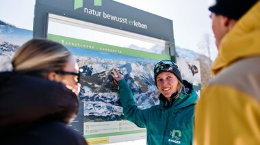 Frau in Winterkleidung zeigt auf ein großes Informationsschild mit Bergbildern, während zwei andere Menschen aufmerksam zuhören, im verschneiten Kleinwalsertal. | © Kleinwalsertal Tourismus eGen • Fotograf: Oliver Farys