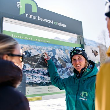 Frau in Winterkleidung zeigt auf ein großes Informationsschild mit Bergbildern, während zwei andere Menschen aufmerksam zuhören, im verschneiten Kleinwalsertal. | © Kleinwalsertal Tourismus eGen • Fotograf: Oliver Farys