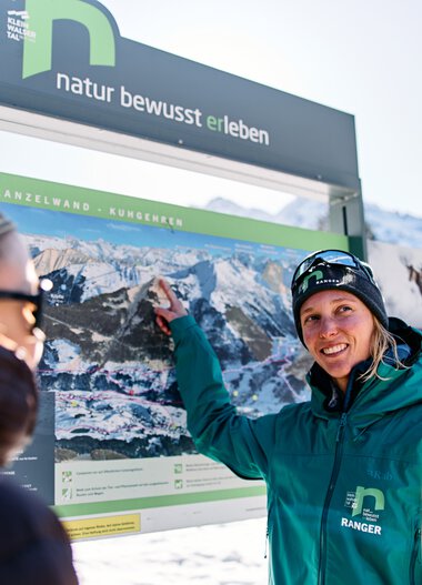 Frau in Winterkleidung zeigt auf ein großes Informationsschild mit Bergbildern, während zwei andere Menschen aufmerksam zuhören, im verschneiten Kleinwalsertal. | © Kleinwalsertal Tourismus eGen • Fotograf: Oliver Farys