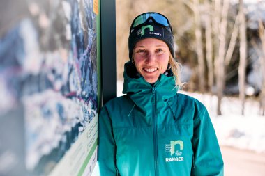 Eine lächelnde Frau in Winterkleidung und mit Skibrille steht neben einem Informationsschild im Schnee, im Hintergrund Bäume und verschneite Landschaft. | © Kleinwalsertal Tourismus eGen • Fotograf: Oliver Farys