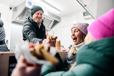 Freundliches Schneemannntreff im Kleinwalsertal, bei dem ein Mann Gästen Essen serviert, während sie lachen und sich freuen. | © Kleinwalsertal Tourismus eGen • Fotograf: Oliver Farys