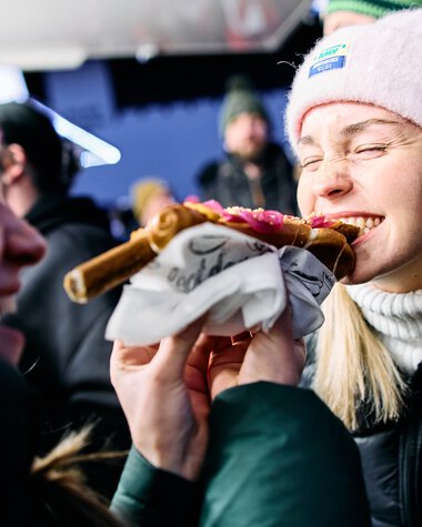 Zwei Frauen beim Schneemanntreff im Kleinwalsertal, eine isst ein Stück Pizza, beide lachen und tragen Winterkleidung. | © Kleinwalsertal Tourismus eGen • Fotograf: Oliver Farys