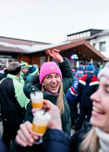 Fröhliche Frau mit pinker Mütze bei Schneemann-Treffen im Kleinwalsertal, umgeben von Menschen bei Winterveranstaltung. | © Kleinwalsertal Tourismus eGen • Fotograf: Oliver Farys