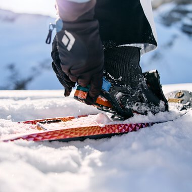 Person snowshoeing in the snow, holding a camera in one hand, with snow-covered mountains in the background. | © Kleinwalsertal Tourismus eGen • Photographer: Oliver Farys