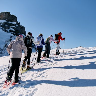 Gruppen von Schneeschuhwanderern auf einem verschneiten Hang in den Bergen, Blick auf alpine Landschaft und Felsen im Hintergrund, sonniges Wetter. | © Kleinwalsertal Tourismus eGen • Fotograf: Oliver Farys
