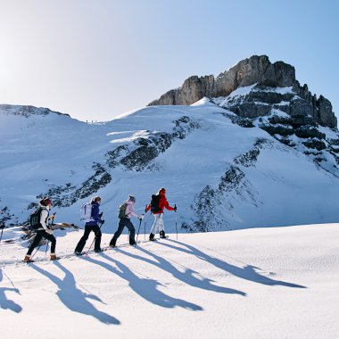 Sechs Personen beim Schneeschuhwandern in verschneiter Berglandschaft im Kleinwalsertal, Sonne am Himmel. | © Kleinwalsertal Tourismus eGen • Fotograf: Oliver Farys