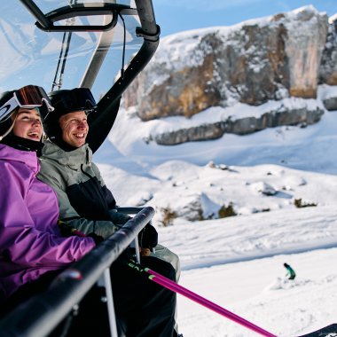 Zwei Personen sitzen im Sessellift im winterlichen Kleinwalsertal, umgeben von schneebedeckter Landschaft und Bergen. | © Kleinwalsertal Tourismus eGen • Fotograf: Oliver Farys