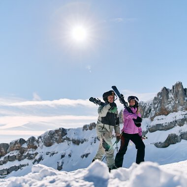 Zwei Skifahrer stehen auf verschneiten Bergen im Sonnenschein, mit einer klaren blauen Himmel und schneebedeckten Gipfeln im Hintergrund. | © Kleinwalsertal Tourismus eGen • Fotograf: Oliver Farys