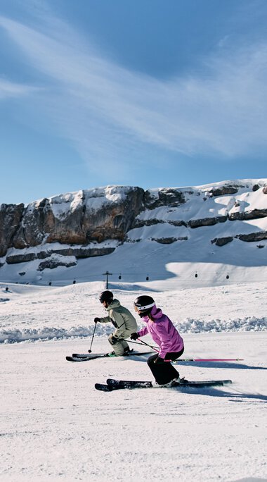 Zwei Kinder beim Skifahren auf winterlicher Piste vor einer Berglandschaft im Kleinwalsertal, mit blauen Himmel und Wolken. | © Kleinwalsertal Tourismus eGen • Fotograf: Oliver Farys