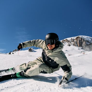 Skifahrer in einem weißen Skianzug trägt einen schwarzen Helm und fährt im Tiefschnee bei klarem Himmel im Skigebiet Ifen. | ©  Kleinwalsertal Tourismus eGen • Fotograf: Oliver Farys