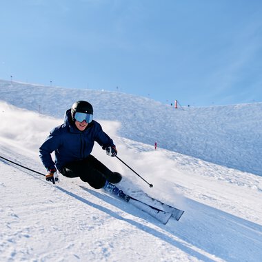Skifahrer in blauer Jacke und dunklen Hosen beim Kurvenfahren auf der schneebedeckten Piste in sonnigem Winterwetter. | © Kleinwalsertal Tourismus eGen • Fotograf: Oliver Farys