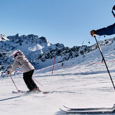 Zwei Skifahrer beim Abfahrtski auf einer verschneiten Piste in den Bergen, im Hintergrund schneebedeckte Gipfel und klarer blauer Himmel. | © Kleinwalsertal Tourismus eGen • Fotograf: Oliver Farys