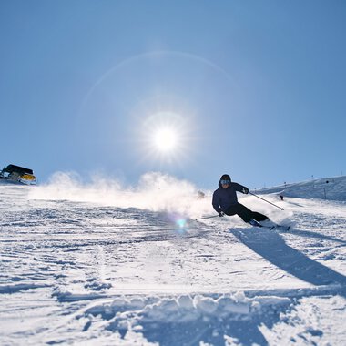Skier descending at Kanzelwand in Kleinwalsertal under a bright sun, with ski lifts and a mountain hut in the background. | © Kleinwalsertal Tourismus eGen • Photographer: Oliver Farys