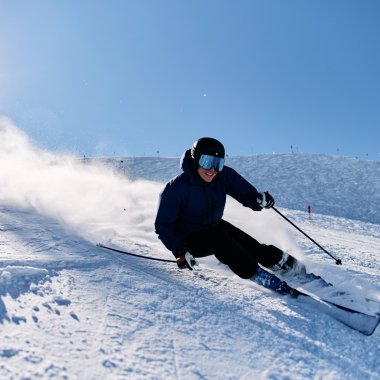 Skifahrer in kniender Position auf einer Schneepiste in Kanzelwand, mit einem blauen Himmel und Sonne im Hintergrund, macht eine schnelle Kurve. | © Kleinwalsertal Tourismus eGen • Fotograf: Oliver Farys
