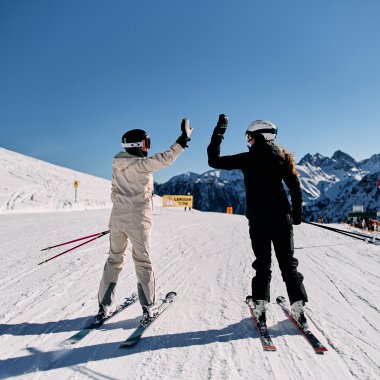Zweimal Skifahrer geben sich auf einer schneebedeckten Piste beim Skifahren in den Bergen ein High-Five, im Hintergrund die Alpen im klaren blauen Himmel. | © Kleinwalsertal Tourismus eGen • Fotograf: Oliver Farys