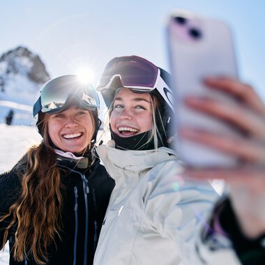 Two smiling women skiers taking a selfie with a smartphone on Kanzelwand in Kleinwalsertal during winter. | © Kleinwalsertal Tourismus eGen • Photographer: Oliver Farys