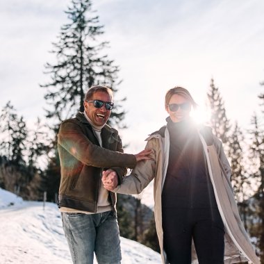 Zwei lachende Menschen auf einer Winterwanderung im Kleinwalsertal, die sich herzlich die Hand geben, umgeben von verschneiten Bäumen. | © Kleinwalsertal Tourismus eGen • Fotograf: Oliver Farys