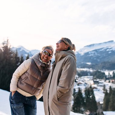 Zwei lachende Frauen genießen eine Winterwanderung in einer verschneiten Landschaft mit Bergen im Hintergrund. | © Kleinwalsertal Tourismus eGen • Fotograf: Oliver Farys