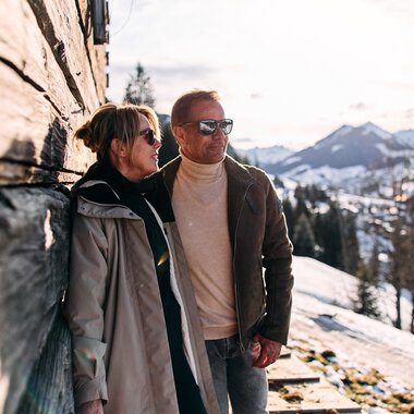 Zwei Menschen stehen bei einer Winterwanderung im Schnee, mit Bergen im Hintergrund, an einer Holzfassade an einem sonnigen Tag. | © Kleinwalsertal Tourismus eGen • Fotograf: Oliver Farys