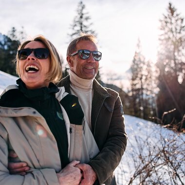 Ein lachendes Paar trägt Sonnenbrillen und genießt eine Winterwanderung in verschneiter Landschaft mit Bergen und Bäumen im Hintergrund. | © Kleinwalsertal Tourismus eGen • Fotograf: Oliver Farys