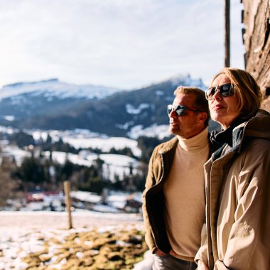 Zwei Personen stehen im Winter in den Bergen, lehnen an einer Holzwand und tragen Sonnenbrillen. Schneebedeckte Berge sind im Hintergrund sichtbar. | © Kleinwalsertal Tourismus eGen • Fotograf: Oliver Farys