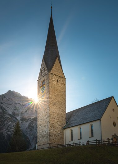 Eine weiße Kirche mit steinernem Turm vor einer Bergkulisse, Sonnenlicht hinter dem Turm, blauer Himmel. | © Kleinwalsertal Tourismus eGen • Fotograf: Robert Niederwolfsgruber