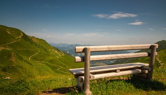 Eine Holzbank auf einer grünen Wiese in den Bergen mit Blick auf den Talboden und sattgrünen Hängen. | © Kleinwalsertal Tourismus eGen • Fotograf: Robert Niederwolfsgruber