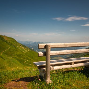 Eine Holzbank auf einer grünen Wiese in den Bergen mit Blick auf den Talboden und sattgrünen Hängen. | © Kleinwalsertal Tourismus eGen • Fotograf: Robert Niederwolfsgruber