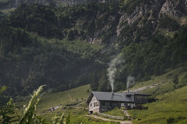 Landschaft mit grünen Hügeln, einer kleinen Hütte mit Rauch aus den Schornsteinen, umgeben von Bergen und Wolken am Himmel. | © Kleinwalsertal Tourismus eGen • Fotograf: Robert Niederwolfsgruber