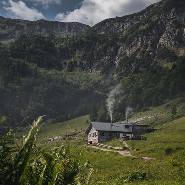 Landschaft mit grünen Hügeln, einer kleinen Hütte mit Rauch aus den Schornsteinen, umgeben von Bergen und Wolken am Himmel. | © Kleinwalsertal Tourismus eGen • Fotograf: Robert Niederwolfsgruber