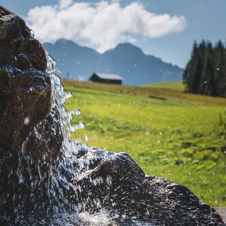 © Kleinwalsertal Tourismus eGen • Fotograf: Robert Niederwolfsgruber Nahaufnahme eines Wasserfalls oder eines fließenden Bachs in einer grünen, bergigen Landschaft mit Wolken und einem kleinen Haus im Hintergrund. | © Kleinwalsertal Tourismus eGen • Fotograf: Robert Niederwolfsgruber
