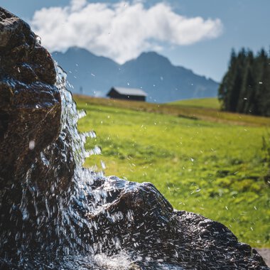 Nahaufnahme eines Wasserfalls oder eines fließenden Bachs in einer grünen, bergigen Landschaft mit Wolken und einem kleinen Haus im Hintergrund. | © Kleinwalsertal Tourismus eGen • Fotograf: Robert Niederwolfsgruber