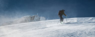 Ein Skifahrer in winterlicher Berglandschaft, der durch den Schnee fährt, bei starkem Wind und hoher Schneeverwehung. | © Kleinwalsertal Tourismus eGen • Fotograf: Robert Niederwolfsgruber