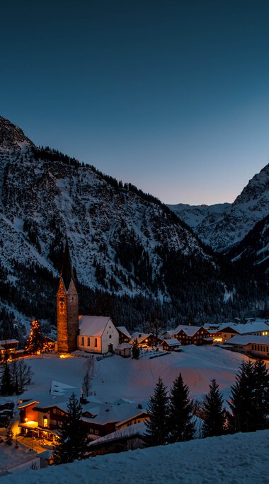 Winterliche Berglandschaft mit einem kleinen Dorf, das in der Nacht in warmem Licht erstrahlt, umgeben von schneebedeckten Bergen. | © Kleinwalsertal Tourismus eGen • Fotograf: Robert Niederwolfsgruber