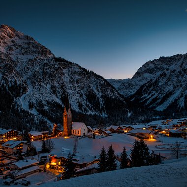 Winterliche Berglandschaft mit einem kleinen Dorf, das in der Nacht in warmem Licht erstrahlt, umgeben von schneebedeckten Bergen. | © Kleinwalsertal Tourismus eGen • Fotograf: Robert Niederwolfsgruber