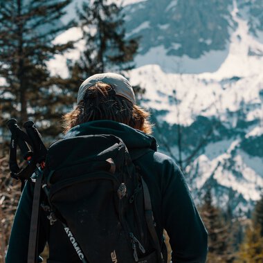 Person mit Rucksack und Wanderstock blickt auf verschneite Berge und Tannenbäume in einer Berglandschaft. | © Kleinwalsertal Tourismus eGen • Fotograf: Distillery