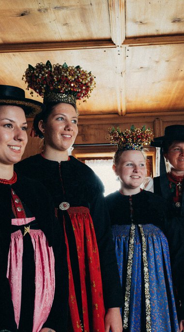 Sechs Frauen in traditioneller bayerischer Tracht stehen nebeneinander in einem Holzraum, lächeln und schauen in eine Richtung. | © Kleinwalsertal Tourismus eGen • Fotograf: Stefan Klauser 