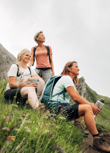 Drei Frauen ruhen sich während einer Wanderung auf einer grünen Bergwiese mit Steinen und Blumen aus, Blick auf die umliegenden Berge. | © Kleinwalsertal Tourismus eGen • Fotograf: Stefan Klauser 