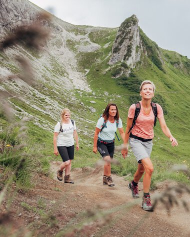Drei Frauen wandern auf einem Pfad in einer grünen Berglandschaft mit steilen Felsen im Hintergrund. | © Kleinwalsertal Tourismus eGen • Fotograf: Stefan Klauser 