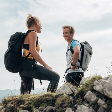 Zwei Wanderer auf einer Bergwanderung, stehend auf einem felsigen Pfad mit Bergblick im Hintergrund. | © Kleinwalsertal Tourismus eGen • Fotograf: Stefan Klauser 