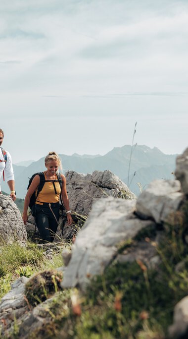 Zwei Wanderer, ein Mann und eine Frau, gehen auf einem Bergpfad mit Felsen und grünem Gras in den Bergen. Das Wetter ist bewölkt.
