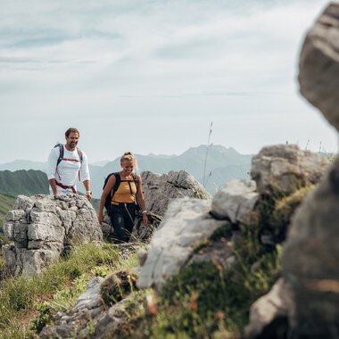 A man and a woman hiking on a mountain trail with rocks and green grass in the mountains. The weather is cloudy.