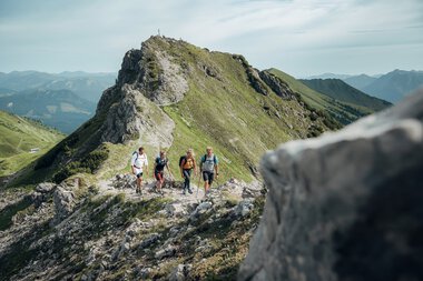 Vier Wanderer auf einem Bergpfad mit Blick auf die Kanzelwand in den Alpen, umgeben von grünen Hängen und Bergen im Hintergrund. | © Kleinwalsertal Tourismus eGen • Fotograf: Stefan Klauser 
