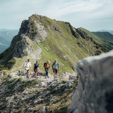 Vier Wanderer auf einem Bergpfad mit Blick auf die Kanzelwand in den Alpen, umgeben von grünen Hängen und Bergen im Hintergrund. | © Kleinwalsertal Tourismus eGen • Fotograf: Stefan Klauser 