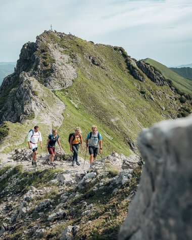 Vier Wanderer auf einem Bergpfad mit Blick auf die Kanzelwand in den Alpen, umgeben von grünen Hängen und Bergen im Hintergrund. | © Kleinwalsertal Tourismus eGen • Fotograf: Stefan Klauser 