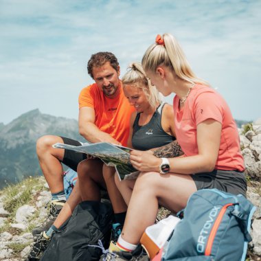 Drei Wanderer, zwei Frauen und ein Mann, sitzen auf einem Felsen und schauen gemeinsam in eine Karte, mit einer Berglandschaft im Hintergrund. | © Kleinwalsertal Tourismus eGen • Fotograf: Stefan Klauser 