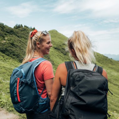 Zwei Frauen mit Rucksäcken wandern in den Bergen, die Landschaft ist grün und bergig. | © Kleinwalsertal Tourismus eGen • Fotograf: Stefan Klauser 